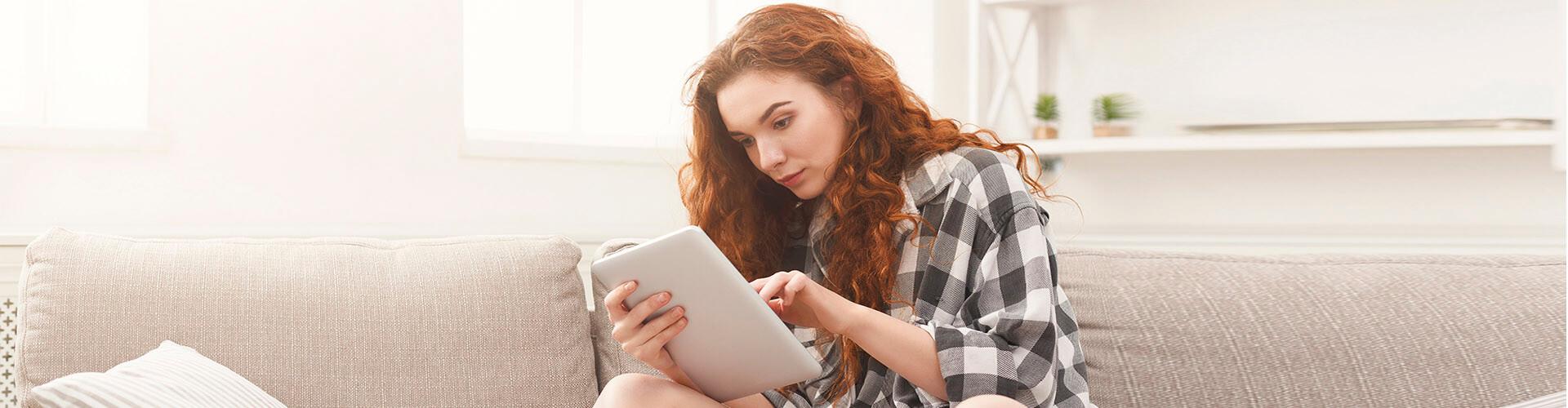 Compte de Pagament Bàsic - Chica jove amb una camisa de quadres asseguda en el sofà de la seua casa amb una tablet